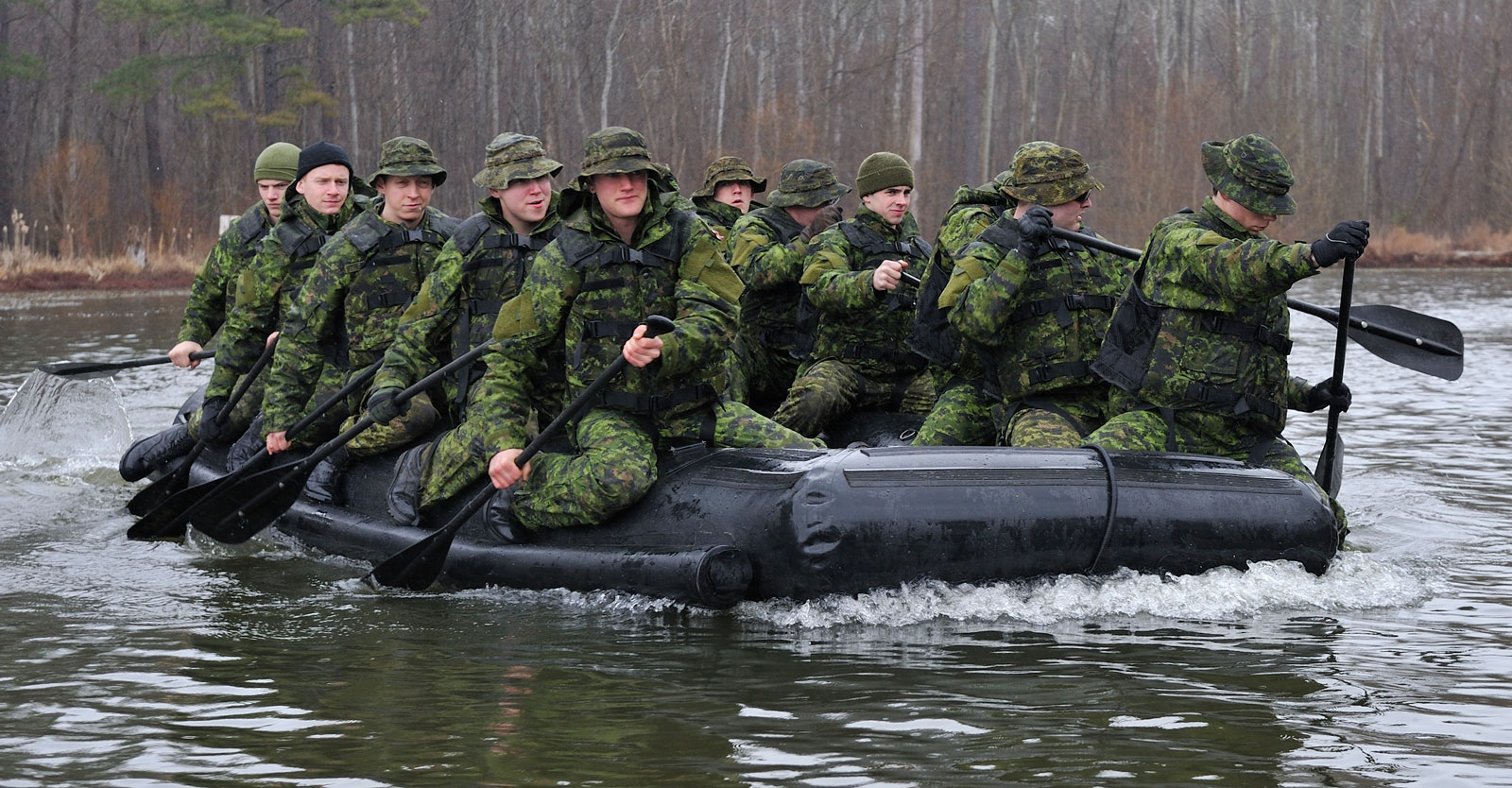 A group of soldiers in camouflage uniforms paddling together in an inflatable raft on a calm river, surrounded by leafless trees, conveying teamwork and focus.
